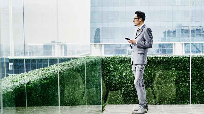 A man in a business suit looks ahead while holding his phone in a sleek glass corridor, with a hedge outside